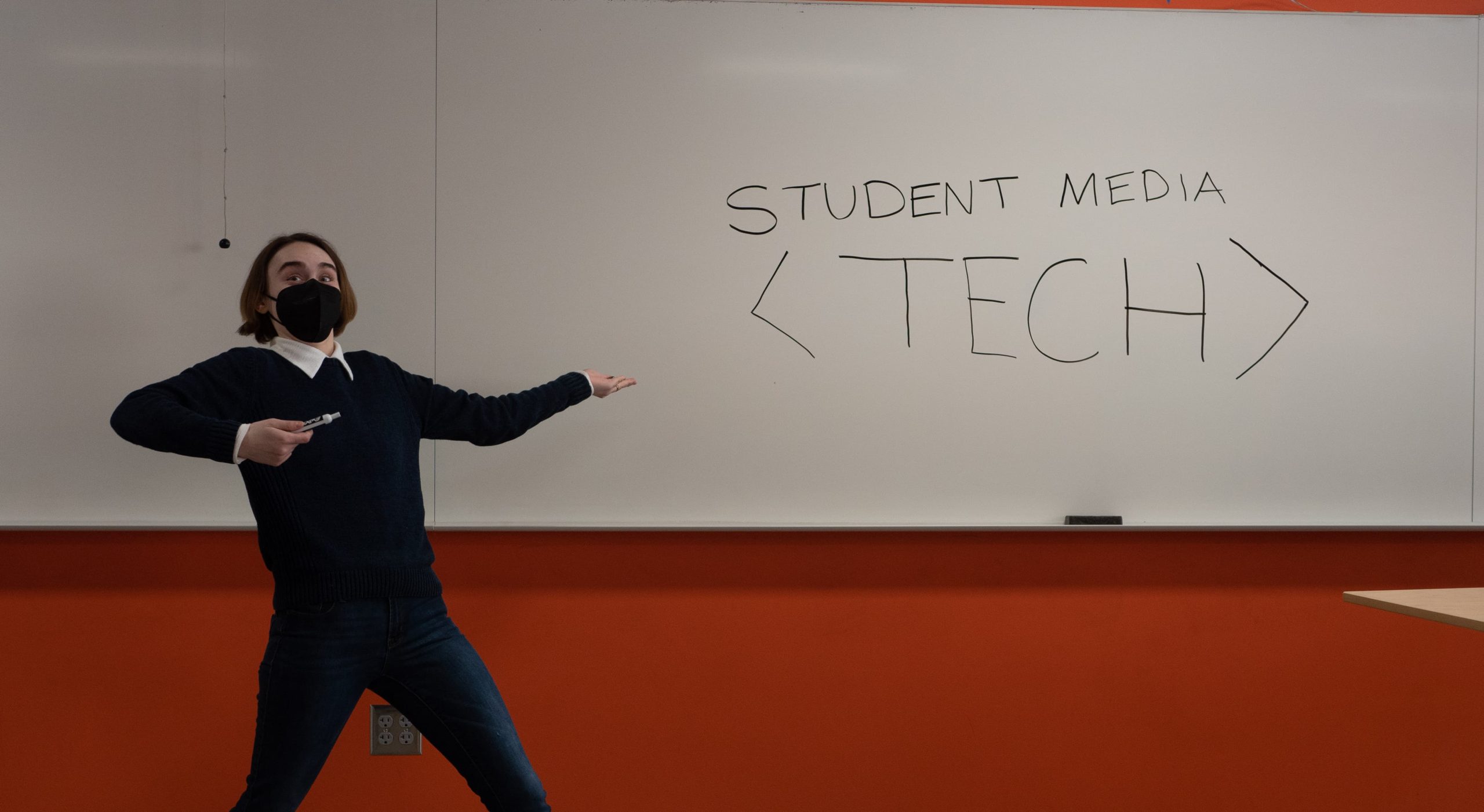 a photo of a person pointing at a whiteboard that reads "student media tech." The photo cuts off at the persons hip level